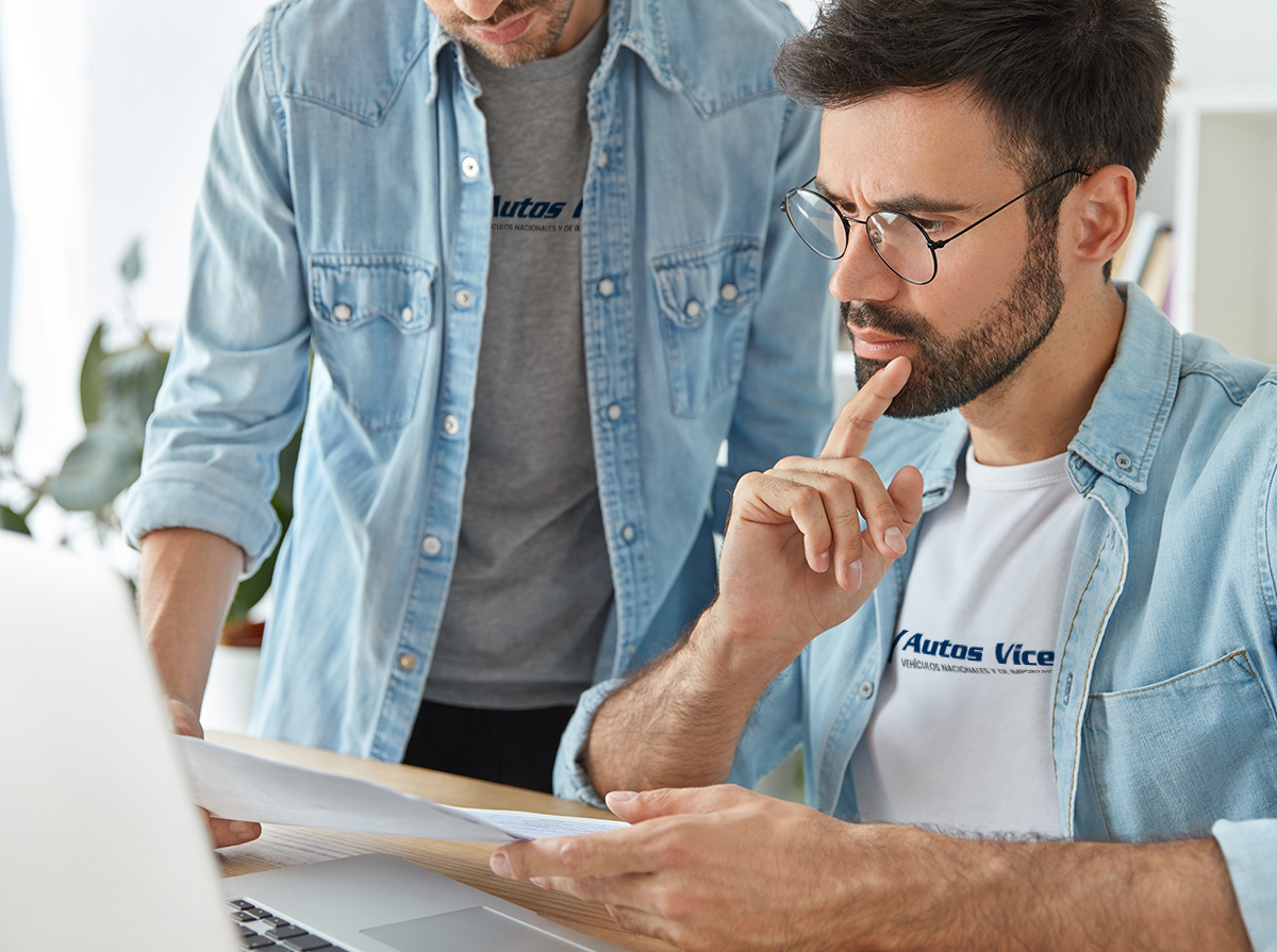 Dos hombres observan un ordenador con un papel en la mano llevando una camiseta con el logotipo de Autos Vicente